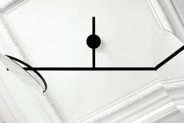 black steel sculpture, with large round mirrors at either end, hanging from white ceiling with ornate plaster mouldings 