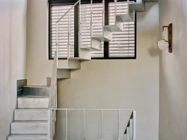Christopher Boots brass wall light hung in an oblique metal stairwell in front of a large, softly lit window 