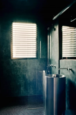 bathroom with dark blue patinated walls and shadowy corners in stark contrast to a stainless steel pedestal sink and neon light 