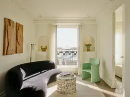 Parisian apartment living room painted off white with a black velvet sofa, a mint green chair, natural textile rug, sculptural wooden wall hangings and featuring a view over the Parisian rooftops 