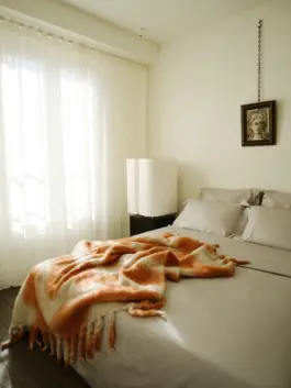 Bedroom with dappled light coming through drapes onto silk sheeted bed with a contemporary orange mohair throw. 